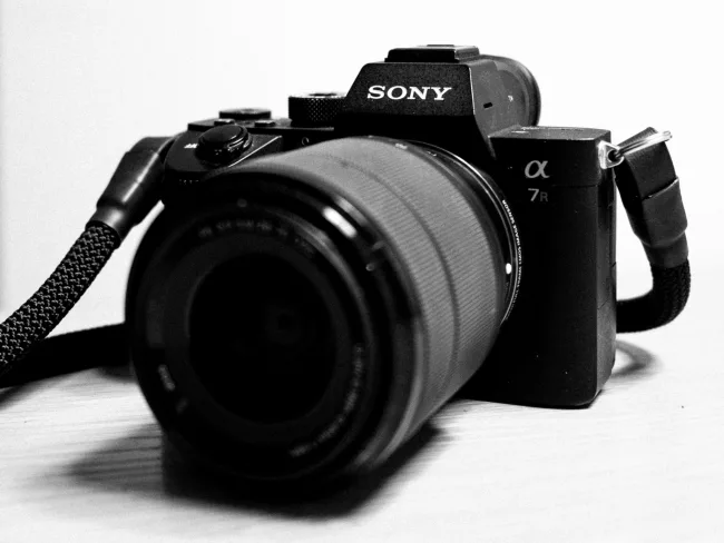 Eine Kamera, genauer, eine Sony A7R iii, liegt auf einer weißen Oberfläche, Es ist ein schwarz-weiß Bild, das Objektiv schaut nach vorne.